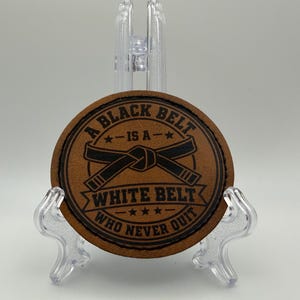 May include: A circular brown leather patch featuring a black engraved graphic of a martial arts belt tied in a knot. The text reads A BLACK BELT IS A WHITE BELT WHO NEVER QUIT, with stars flanking the text and a banner design around the belt graphic.