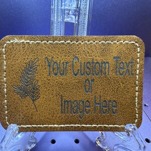 Peut inclure: Un écusson rectangulaire en cuir beige aux coins arrondis avec des coutures claires sur le pourtour. L'écusson présente une gravure de feuille de fougère sur la gauche et le texte "Your Custom Text or Image Here" gravé sur la droite.