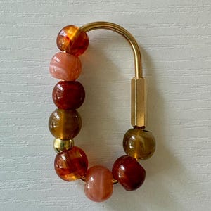 May include: A gold-toned metal carabiner clip adorned with a series of round beads in shades of amber, brown, and marbled peach. The beads are arranged along the curved side of the clip, featuring a mix of translucent and opaque finishes.