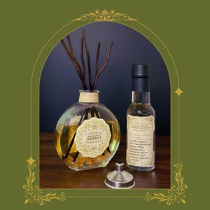 May include: A round glass reed diffuser filled with yellow liquid and orange stones, featuring dark brown reeds and a label reading Bergamot Orange. Beside it stands a clear bottle with a black cap and a label titled The Botanical Conservatory.