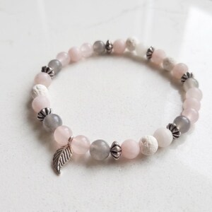 May include: A beaded bracelet featuring alternating light pink, translucent grey, and white porous beads. The bracelet is accented with small, silver-toned metal spacers and a single silver-toned metal leaf charm hanging from the circular strand.