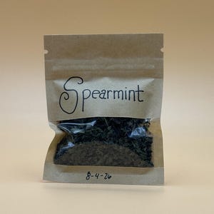 May include: A small, rectangular, brown paper pouch with a clear plastic window containing dried, dark green spearmint leaves. The word "Spearmint" is handwritten in black ink above the window, and the date "8-4-26" is written in black ink below.
