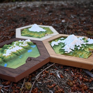 May include: Three hexagonal wooden frames containing 3D topographical mountain maps. Each map features green terrain, blue water elements, and white snow-capped peaks. The frames are made of different wood tones, and one includes the text Blazing Goat.