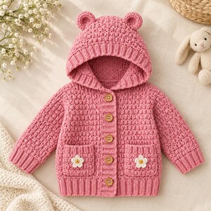 May include: A pink long-sleeve hooded cardigan with a textured knit pattern. The garment features a hood with two small bear ears, five light brown wooden buttons down the centre, and two front pockets each adorned with a white flower applique.