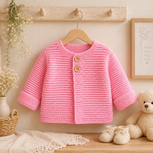 May include: A bright pink long-sleeve knit cardigan featuring a round neckline and two light brown wooden buttons down the centre front. The garment is constructed with a horizontal garter stitch pattern and has folded cuffs at the end of each sleeve.