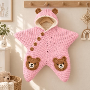 May include: A star-shaped baby bunting bag crocheted in light pink yarn, featuring a hood with two small brown bear ears. The front has four wooden buttons and two brown bear face appliques. The interior is lined with cream-colored fabric.