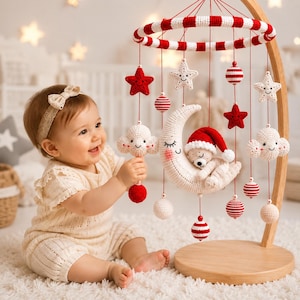 Puede incluir: Un móvil de ganchillo para bebé con soporte de madera y marco circular de rayas rojas y blancas. Los elementos colgantes incluyen una luna creciente con un oso dormido con gorro rojo, nubes sonrientes, estrellas y esferas a rayas rojas y blancas.