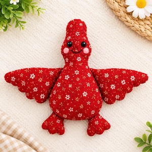 Könnte beinhalten: Ein Plüschvogel-Spielzeug aus rotem Stoff mit weißem Blumenmuster. Der Vogel hat zwei schwarze Perlenaugen, einen kleinen gestickten Schnabel und rosa kreisförmige Flecken auf den Wangen. Er verfügt über ausgebreitete Flügel und zwei Füße aus demselben Stoff.