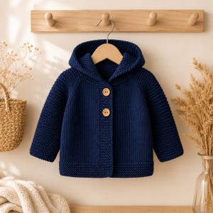 May include: A dark navy blue hooded cardigan made of a thick, textured knit fabric. The garment features long sleeves, a hood, and two large, round, light-coloured wooden buttons centred on the front placket. The knit pattern is consistent throughout.