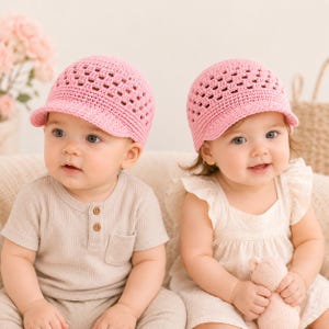 May include: Two identical light pink crochet baseball caps featuring a textured, open-weave pattern on the crown and a solid, tightly stitched brim. The caps are constructed from yarn and designed with a classic rounded shape and a short, curved visor.