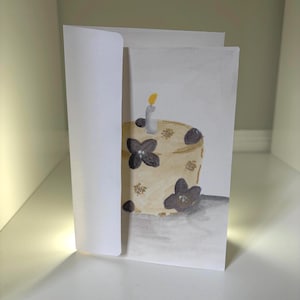 May include: A white greeting card featuring a watercolor illustration of a beige cake decorated with dark purple flowers and gold speckles. A single lit candle with a yellow flame sits on top of the cake, which casts a gray shadow on the surface.