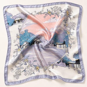 May include: A square silk scarf featuring a lavender border and a central illustration of a landscape with traditional buildings, trees, and a body of water. The design is framed by a floral wreath with yellow and white flowers against a soft pink sky.