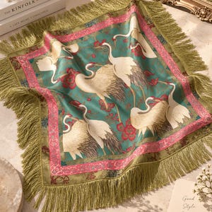 May include: A square fabric mat featuring a teal background with white crane birds and red floral patterns. The mat is bordered by a pink decorative frame and finished with long olive green fringe trim on all four sides, creating a textured edge.