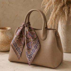 May include: A taupe-colored handbag with a textured surface, featuring two top handles and a decorative square scarf tied around one handle. The scarf displays a detailed paisley pattern in shades of brown, blue, pink, and cream with a geometric border.