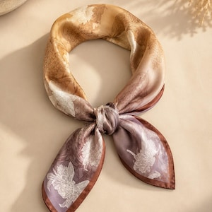 May include: A square silk scarf featuring a gradient color transition from warm golden brown to muted mauve. The fabric is decorated with a white line-art floral pattern and finished with a dark brown border, tied in a simple knot at the center.