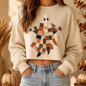 May include: A cream-colored long-sleeve crop sweatshirt featuring a large central ghost graphic composed of a patchwork of various fabric patterns in shades of orange, brown, black, and white. The garment has a crew neckline and ribbed cuffs.