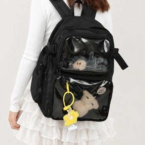 May include: A black backpack featuring two clear front pockets, each containing a small plush toy. The lower pocket displays a yellow flower-shaped keychain and a circular patch with the text MY BAG. The bag has adjustable straps and side buckles.