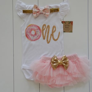 May include: A white bodysuit featuring a donut graphic and gold glitter text reading "ONE". The set includes a pink tutu diaper cover with a gold sequin bow and a matching headband with a bow.