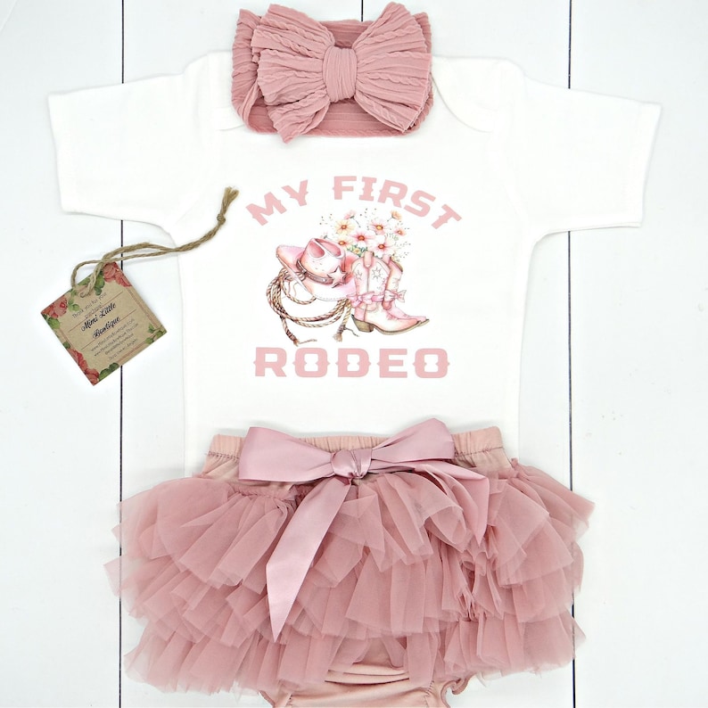 May include: A mauve chiffon bloomers, a white onesie with a mauve pink text "My first rodeo ", a mauve pink nylon bow headband.