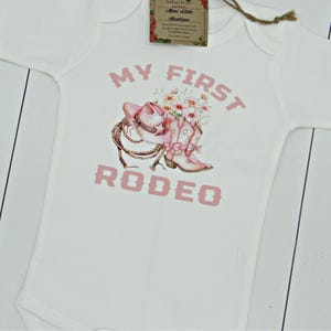 May include: White baby onesie with pink text that reads "My First Rodeo". A pink cowboy hat, boots, and flowers are illustrated on the onesie.