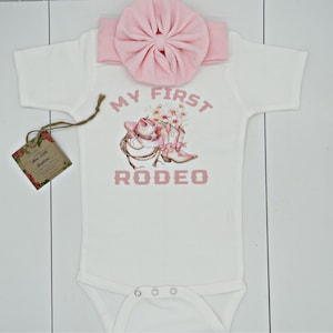 May include: A white baby onesie with pink text that reads "My First Rodeo". The onesie features a pink cowboy hat and boots with flowers. A pink bow headband is included.