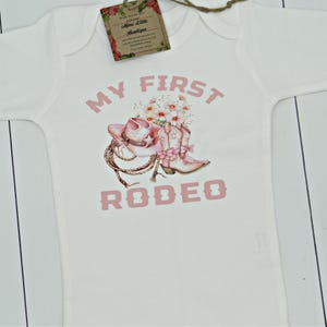 May include: White baby onesie with pink text that reads "My First Rodeo". The onesie features a pink cowboy hat and boots with flowers.