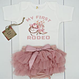 May include: A white baby onesie with pink text that reads "My First Rodeo". The onesie has a pink graphic of a cowboy hat, boots, and flowers. The onesie is paired with a pink tulle skirt with a large pink satin bow.
