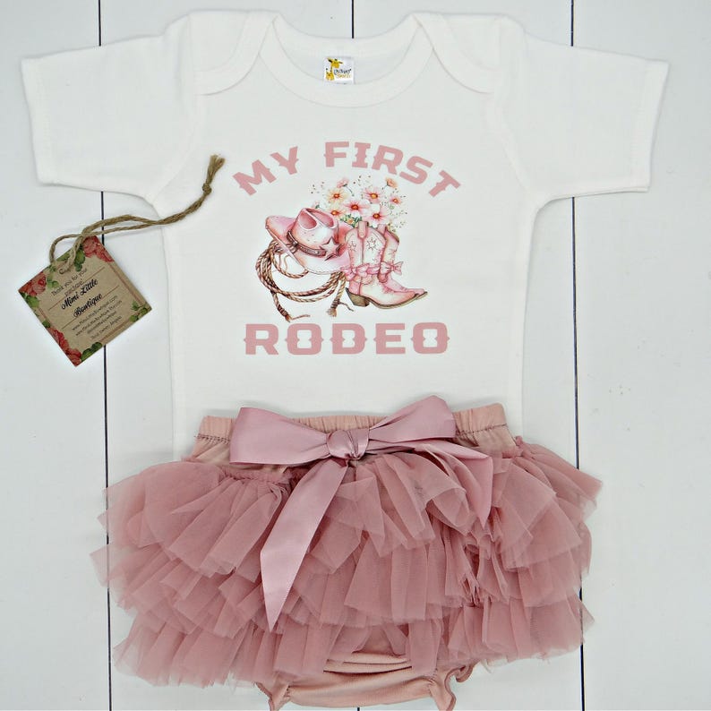 May include: A white baby onesie with pink text that reads "My First Rodeo". The onesie has a pink graphic of a cowboy hat, boots, and flowers. The onesie is paired with a pink tulle skirt with a large pink satin bow.