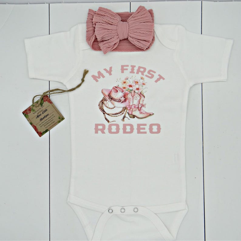 May include: White baby onesie with pink text that reads "My First Rodeo". The onesie features a pink cowboy hat with flowers and pink cowboy boots.