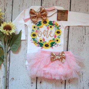 May include: A girl's first birthday outfit with a white long-sleeved bodysuit featuring a sunflower wreath with the word "one" in gold glitter, a pink tulle tutu skirt, and  gold glitter bow.