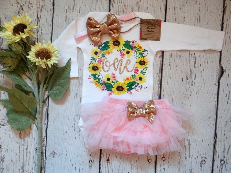 May include: A girl's first birthday outfit with a white long-sleeved bodysuit featuring a sunflower wreath with the word "one" in gold glitter, a pink tulle tutu skirt, and  gold glitter bow.