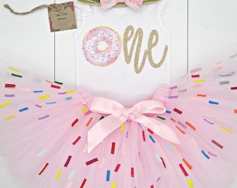 May include: A white bodysuit with a donut graphic and gold glitter "ONE" text, paired with a pink tutu skirt decorated with colorful confetti. A matching pink bow headband and a gold glitter headband complete the set. A tag is attached.