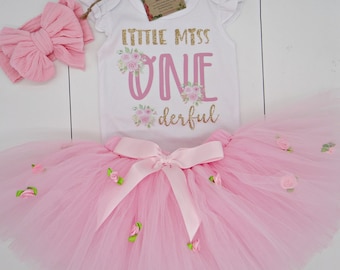 May include: A pink tutu with a pink bow and floral accents, a white onesie with the text "Little Miss One-derful" in pink and gold glitter, and a pink headband.