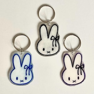 Puede incluir: Tres llaveros con forma de conejo y anillas de metal plateado. Cada llavero presenta una cabeza de conejo blanca translúcida con dos orejas largas y un pequeño lazo. Los contornos están delineados en colores azul, negro y morado.