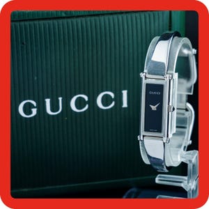 May include: A silver-toned Gucci wristwatch featuring a rectangular black dial with silver hands and the Gucci logo. The watch has a polished metal bangle-style band and is displayed in front of a dark green box with the white Gucci brand name.