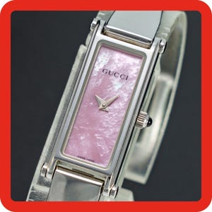 Könnte beinhalten: Eine rechteckige Gucci-Armbanduhr mit einem polierten silberfarbenen Metallgehäuse und einem strukturierten hellrosa Perlmuttzifferblatt. Die Uhr hat silberfarbene Zeiger, eine seitliche Krone mit dunklem Akzent sowie den Schriftzug GUCCI und SWISS MADE.