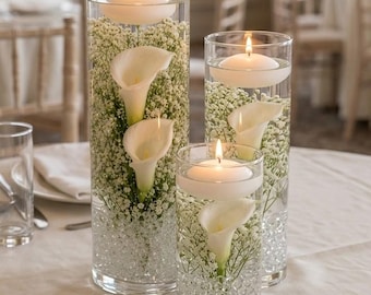 May include: Three clear cylindrical glass vases filled with water, small clear beads, white baby's breath flowers, and white calla lilies. Each vase is topped with a single white floating candle, creating a tiered arrangement of floral centerpieces.