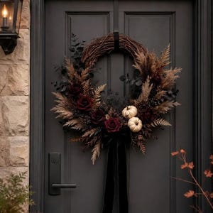 May include: A circular wreath made of brown twigs, dark red roses, dried pampas grass, and dark green foliage. Two small white pumpkins are nestled within the arrangement, which is accented by a long black velvet ribbon hanging from the bottom.