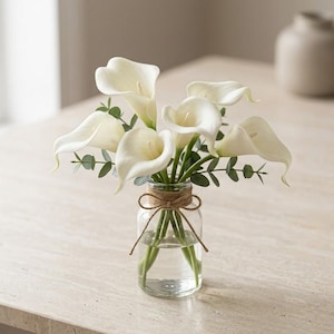 May include: A bouquet of white calla lilies with green eucalyptus leaves arranged in a clear glass jar. The jar is tied with a thin brown twine bow around the neck, holding the stems upright in a simple, clean display of floral decor.