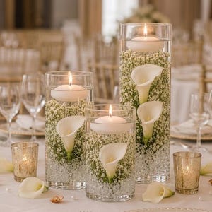 May include: Three clear cylindrical glass vases filled with water, white baby's breath flowers, and submerged white calla lilies. Each vase is topped with a white floating candle. Two small gold mercury glass votive candle holders sit nearby.