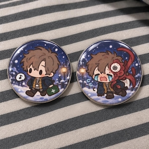 May include: Two circular enamel pins featuring a cartoon character in a snowy setting. The left pin shows the character walking with a green medical kit. The right pin depicts the same character crying with a red, tentacled creature attached.