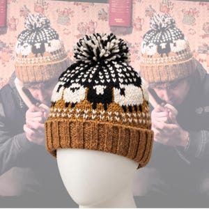 May include: A knit beanie featuring a repeating pattern of white and black sheep against a tan background. The hat has a ribbed cuff, a fair isle style band with sheep motifs, and a large, multi-colored pom-pom on the top in black, white, and tan.