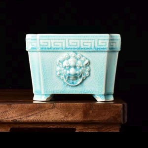 May include: A light blue ceramic planter with a crackle glaze finish. The rectangular vessel features a relief sculpture of a lion head holding a ring on the front panel and a geometric Greek key pattern along the top rim, supported by four feet.
