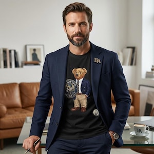 Könnte beinhalten: Ein marineblaues Sakko über einem schwarzen T-Shirt mit einem Teddybären-Aufdruck, der ein blaues Sakko, eine beige Hose und eine Brille trägt. Der Bär steht neben einem runden Logo mit der Aufschrift Brooklyn NY. Eine goldene BA-Anstecknadel am Revers.