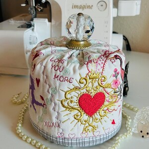 May include: A white fabric pincushion with embroidered details, including a red heart, floral accents, and the words "LOVE YOU MORE." The pincushion has a crystal knob and is surrounded by a pearl necklace.