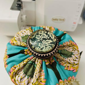 May include: A teal fabric pincushion with a floral design in yellow, pink, and green, topped with a decorative button featuring a black and white floral pattern. The pincushion is near a sewing machine.