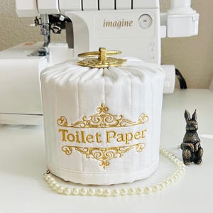 May include: White quilted toilet paper cover with gold embroidered text that reads "Toilet Paper". The cover has a gold handle and is next to a small bronze rabbit figurine and a pearl necklace.