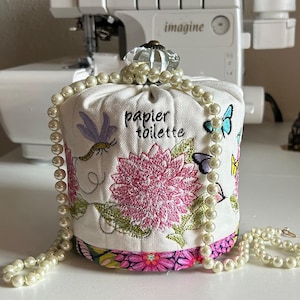 May include: A white fabric toilet paper holder with floral and butterfly embroidery. The holder has the words "papier toilette" embroidered on it. A pearl necklace is draped over the holder.