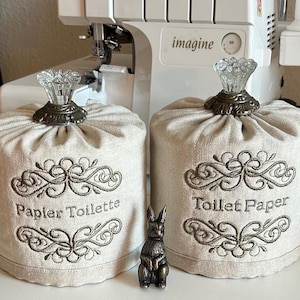 May include: Two beige toilet paper covers with ornate embroidery and crystal-topped finials. One cover reads "Papier Toilette," the other "Toilet Paper." A small bronze rabbit figurine sits between them. The covers are on a white surface.