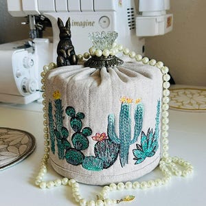 May include: A cylindrical beige fabric cover featuring embroidered green cacti with small yellow and pink flowers. A small bronze-colored rabbit figurine sits on top next to a clear glass finial. A long string of white faux pearls drapes around the item.